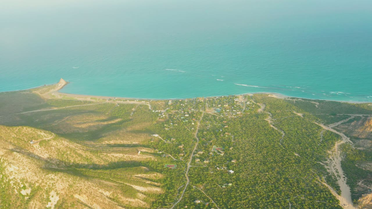 High aerial view of coastal hills, shoreline, turquoise water, and small buildings.