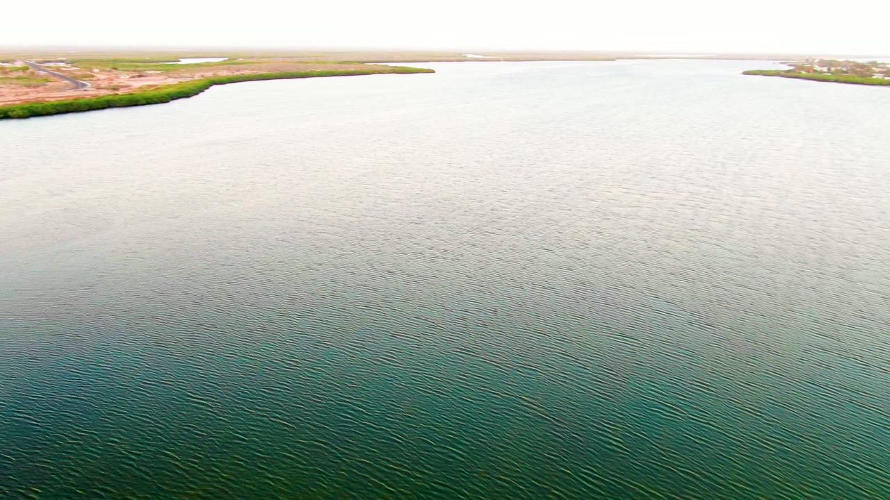 Aerial view in San Carlos, Baja California Sur looking over green-blue rippled water toward a thin vegetated shoreline, dry land, and a pale horizon, with no visible people.