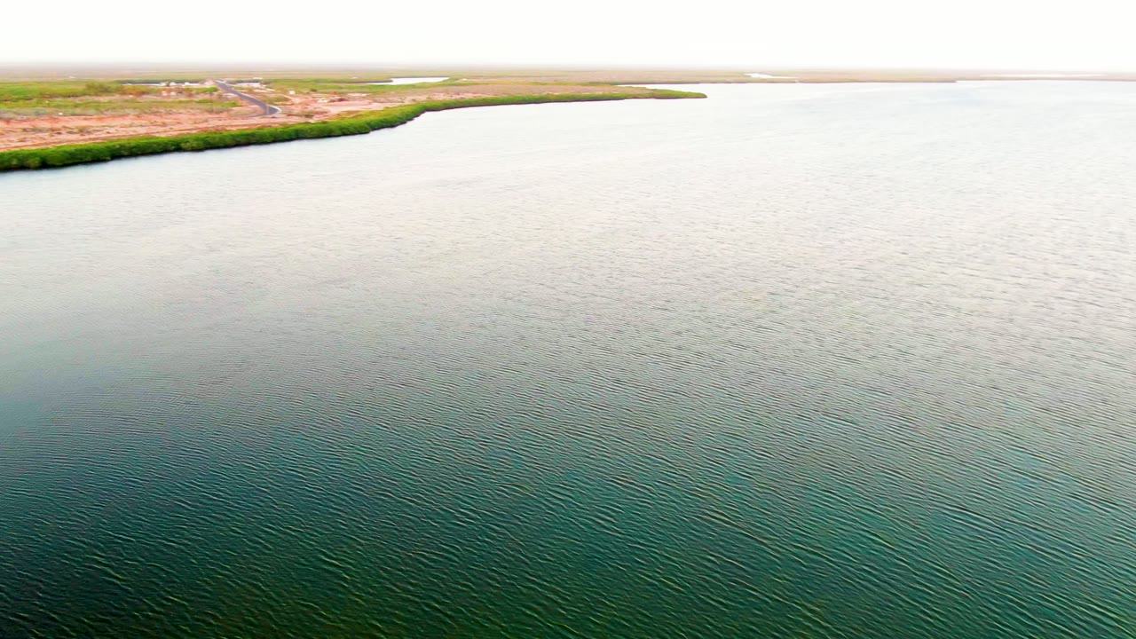 Drone view over calm rippled water in San Carlos, Baja California Sur, with shoreline brush, reddish-brown land, faint structures, and a bright hazy sky, with no visible people.