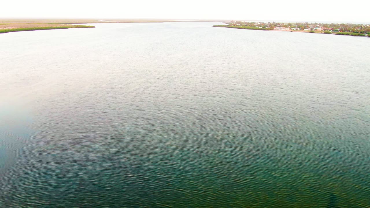 Wide aerial frame in San Carlos showing rippled water in the foreground and midground, shoreline buildings across the upper frame, and no visible people.