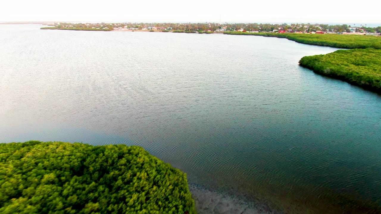 Aerial frame over San Carlos, Baja California Sur showing a wider span of calm water, mangroves at the lower left and right edge, faint sandy shallows near the bottom, distant buildings, and no visible people.