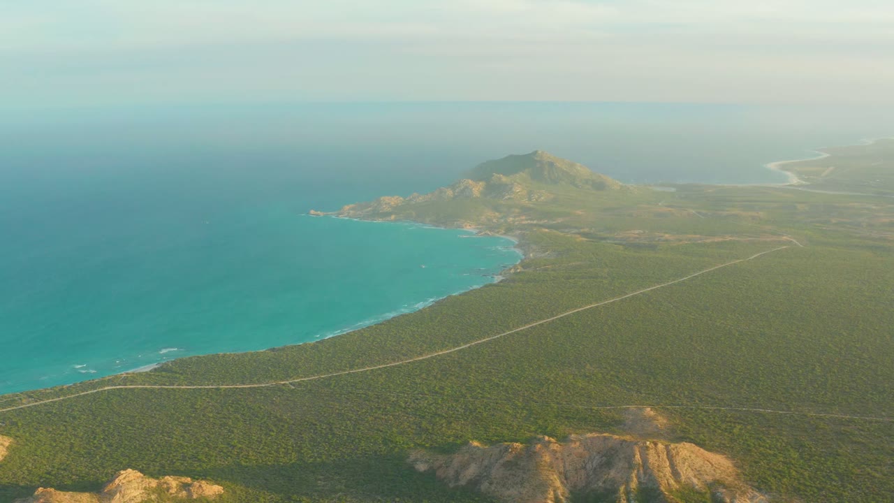 Drone view of coastal road running beside turquoise ocean and green land.