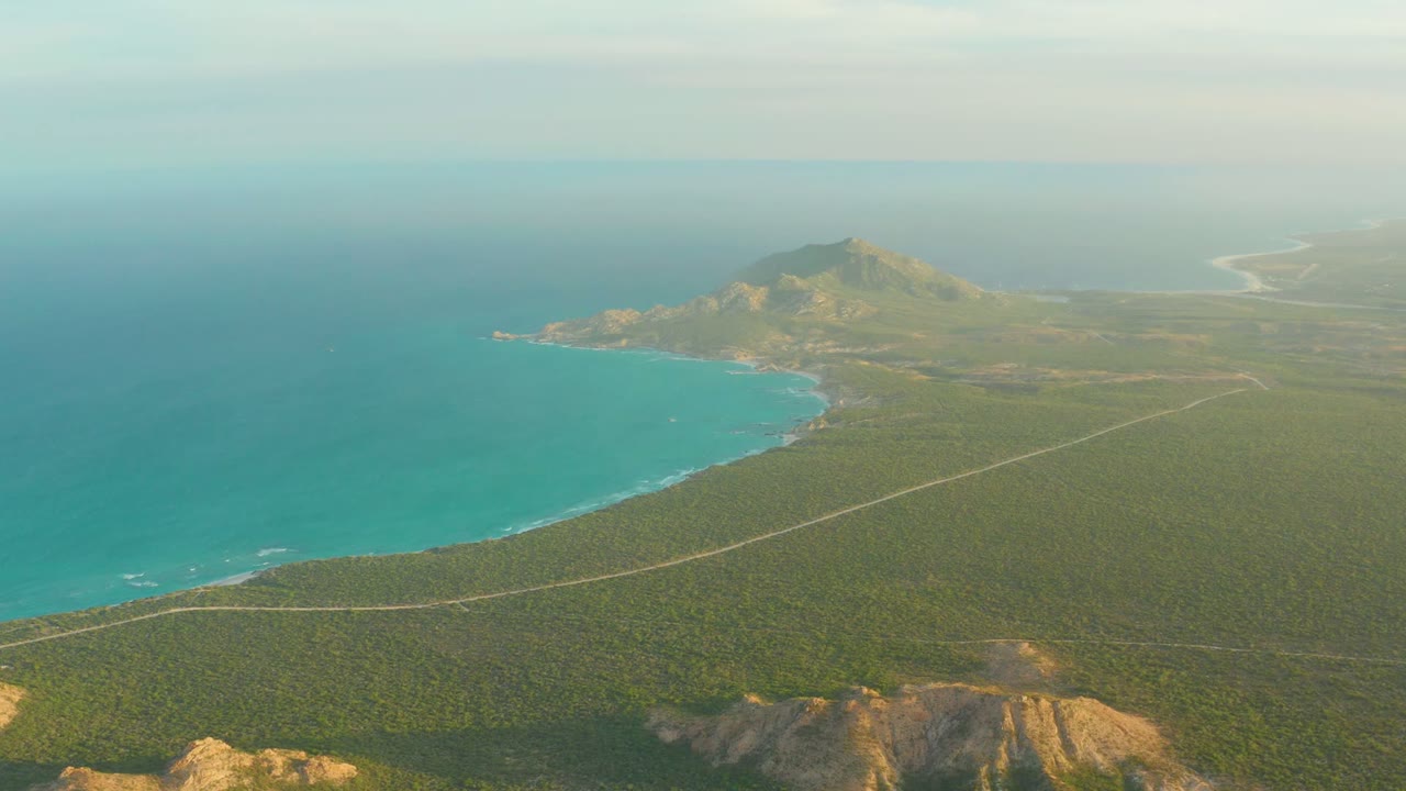 High view over Cabo Pulmo coastline with the ocean filling the left side, the road crossing the lower midground, a prominent hill near the shore, and no visible people.