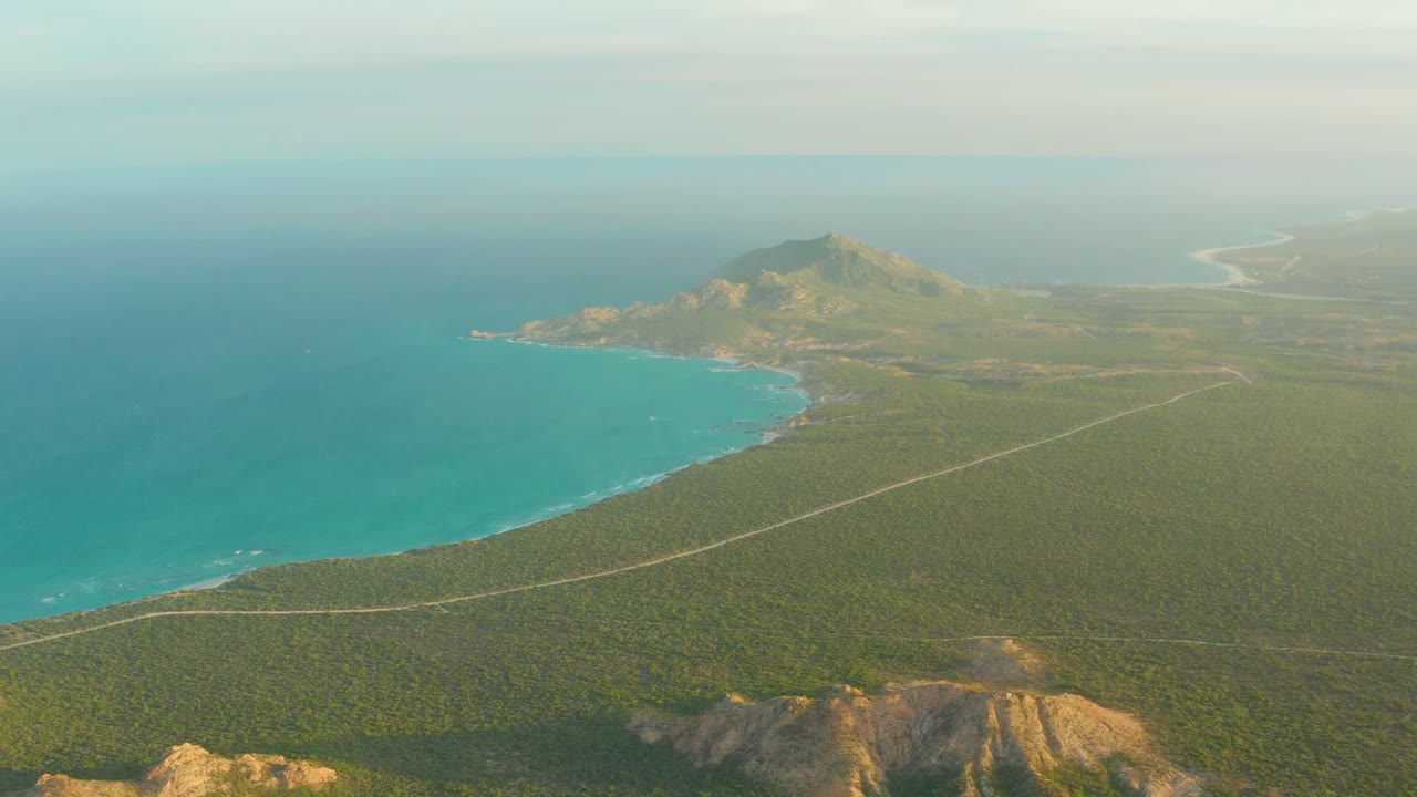 Opening aerial frame over Cabo Pulmo shows turquoise water on the left, a pale coastal road through green terrain, distant hills, hazy sky, and no visible people.