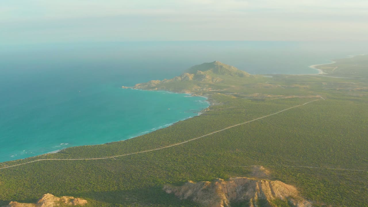At 0:01, the Cabo Pulmo aerial view holds on the curved turquoise bay, the road crossing the green midground, a rocky foreground ridge, and no visible people.