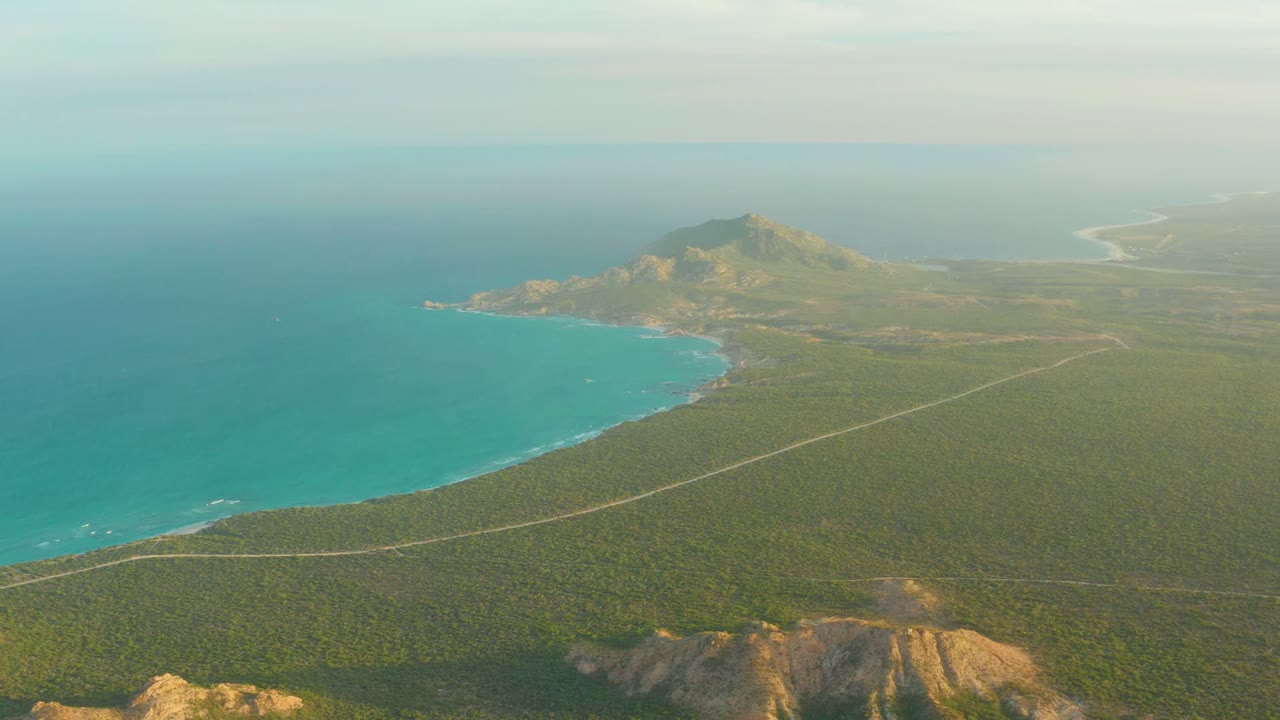 At 0:02, the aerial frame shows Cabo Pulmo shoreline with bright turquoise water, a thin road line over green land, distant coastline, and no visible people.