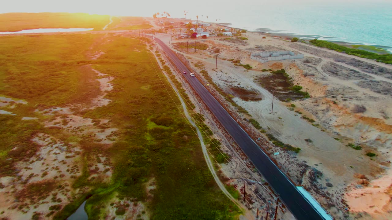 Aerial sunset view over San Carlos, Baja California Sur, with a coastal road running between green grassland, pale dunes, distant structures, and ocean, no visible people.