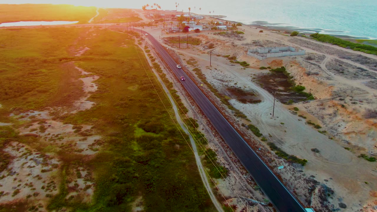 Coastal highway with small vehicles, sandy terrain, grassland, and ocean under warm sun.