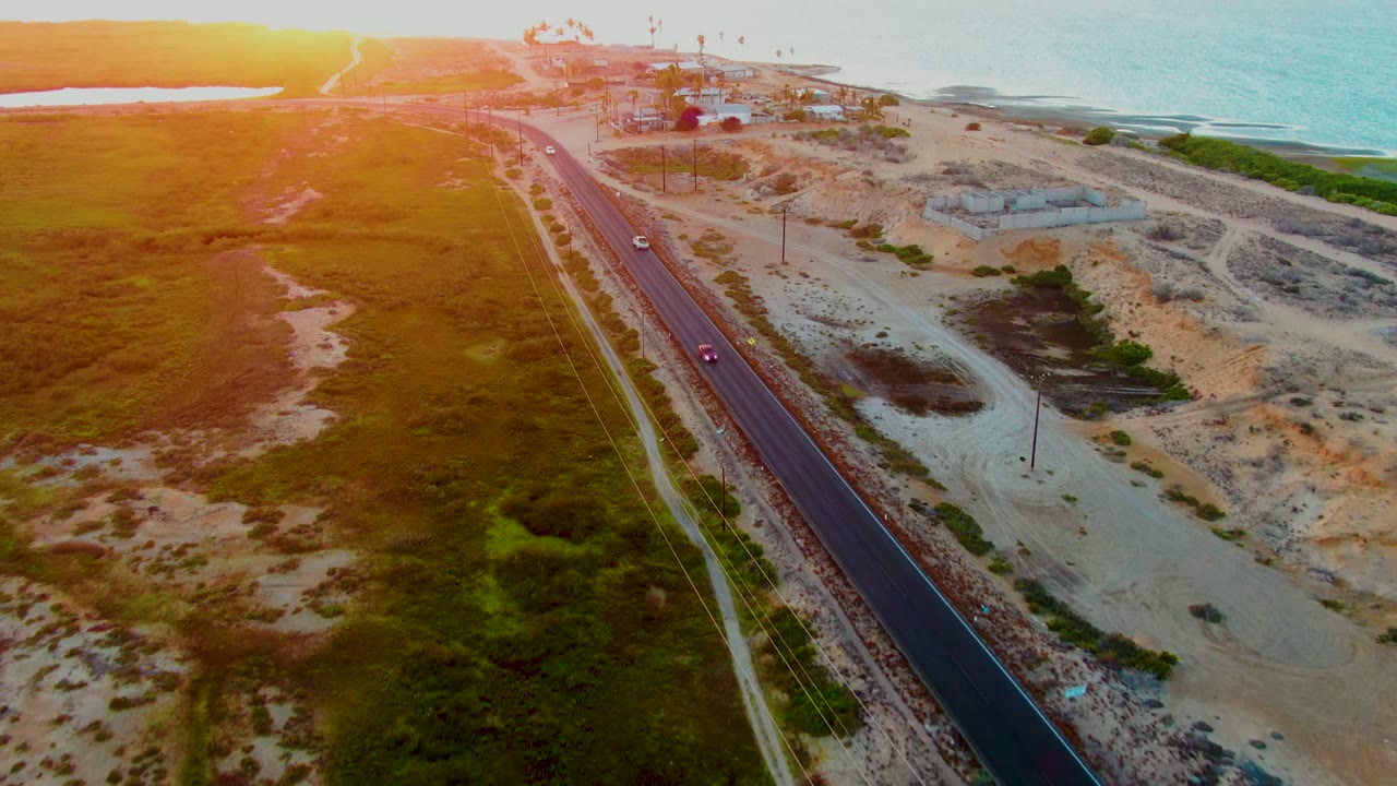 Aerial view in San Carlos with the road leading toward the upper coastline, cars spaced along the asphalt, green marshlike ground on one side, sandy dunes on the other, and no visible people.