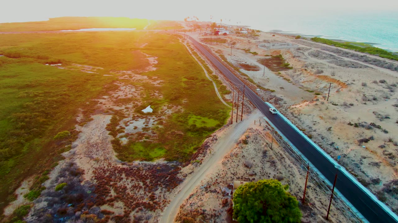 Aerial view in San Carlos, Baja California Sur, with a car on a coastal road between green fields and sandy dunes under bright sunset light, no visible people.