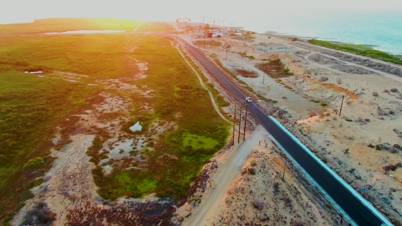 The road angles through San Carlos, Baja California Sur, with the car farther along the lane, sandy dunes to the right, open green terrain to the left, and ocean near the horizon, no visible people.