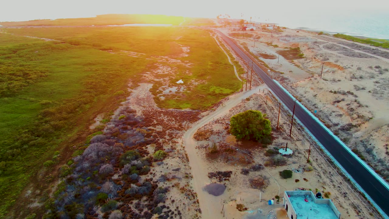 Aerial frame from San Carlos, Baja California Sur showing a dark coastal road between green fields and sandy ground under low sunset light, with no visible people.