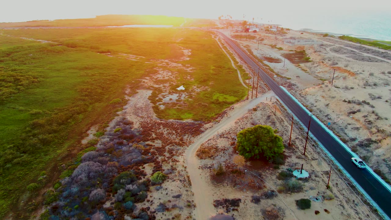 The white car continues along the diagonal paved road in San Carlos, Baja California Sur, passing sandy coastal ground and utility poles with green field texture on the opposite side and no visible people.