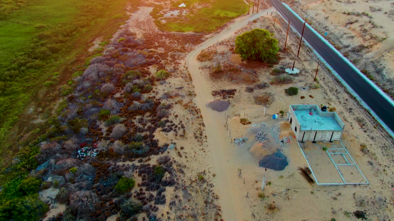 Drone-style overhead view in San Carlos, Baja California Sur with a paved road, utility poles, brushy desert, a small flat-roofed building, and no visible people.