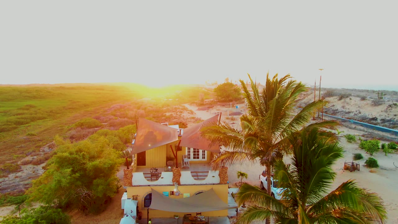 Aerial view in San Carlos, Baja California Sur of a beach house partly covered by palm fronds, with sunset glare over low dunes and no visible people.