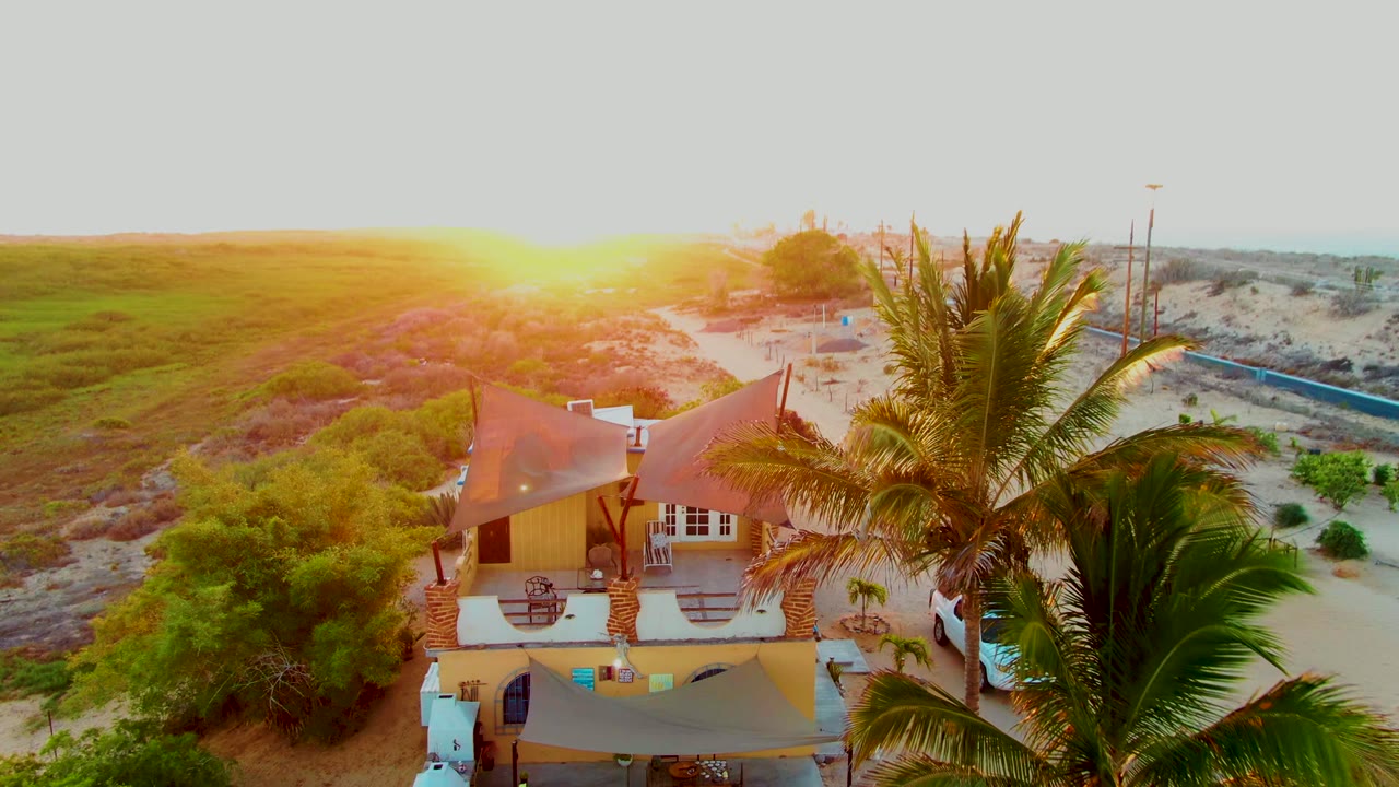 Drone view of a beach house roof, palm tree, sand, and sunset-lit dunes.