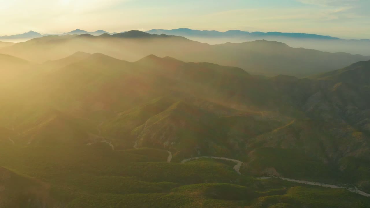 Wide aerial view over Cabo Pulmo mountain valleys at dawn, with warm light entering from the left, layered ridges in haze, a winding pale line in the lower valley, and no visible people.
