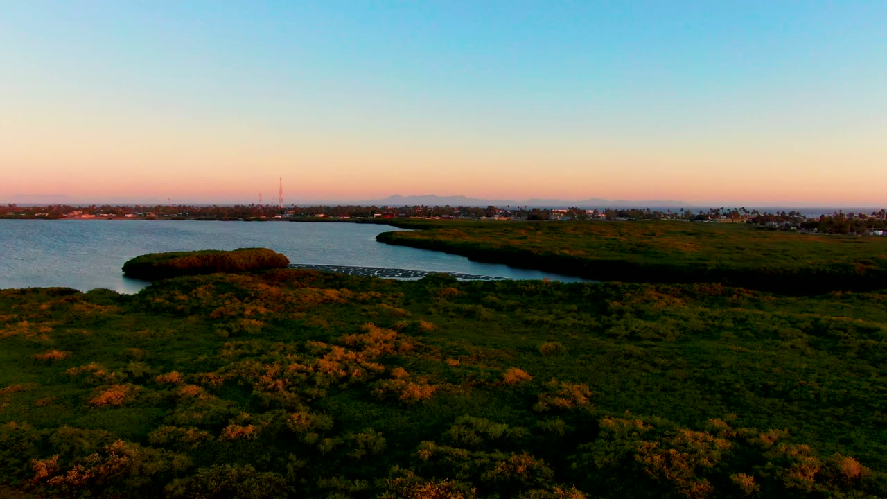 Aerial dusk view in San Carlos, Baja California Sur showing grassy foreground, a curving waterway, a small dock near the left edge, distant shoreline, pink-orange horizon, and no visible people.