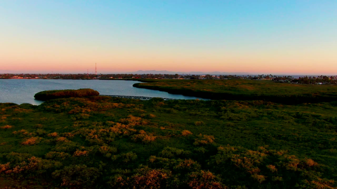 Wide aerial view in San Carlos, Baja California Sur showing darkening shore vegetation, a blue water channel, distant land across the bay, warm sunset color near the horizon, and no visible people.
