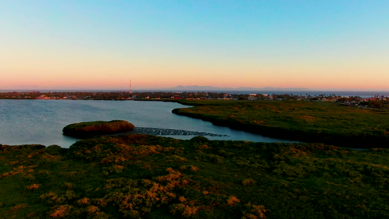 Aerial dusk view of a San Carlos, Baja California Sur inlet bordered by dark green grass, with distant buildings on the horizon and no visible people.