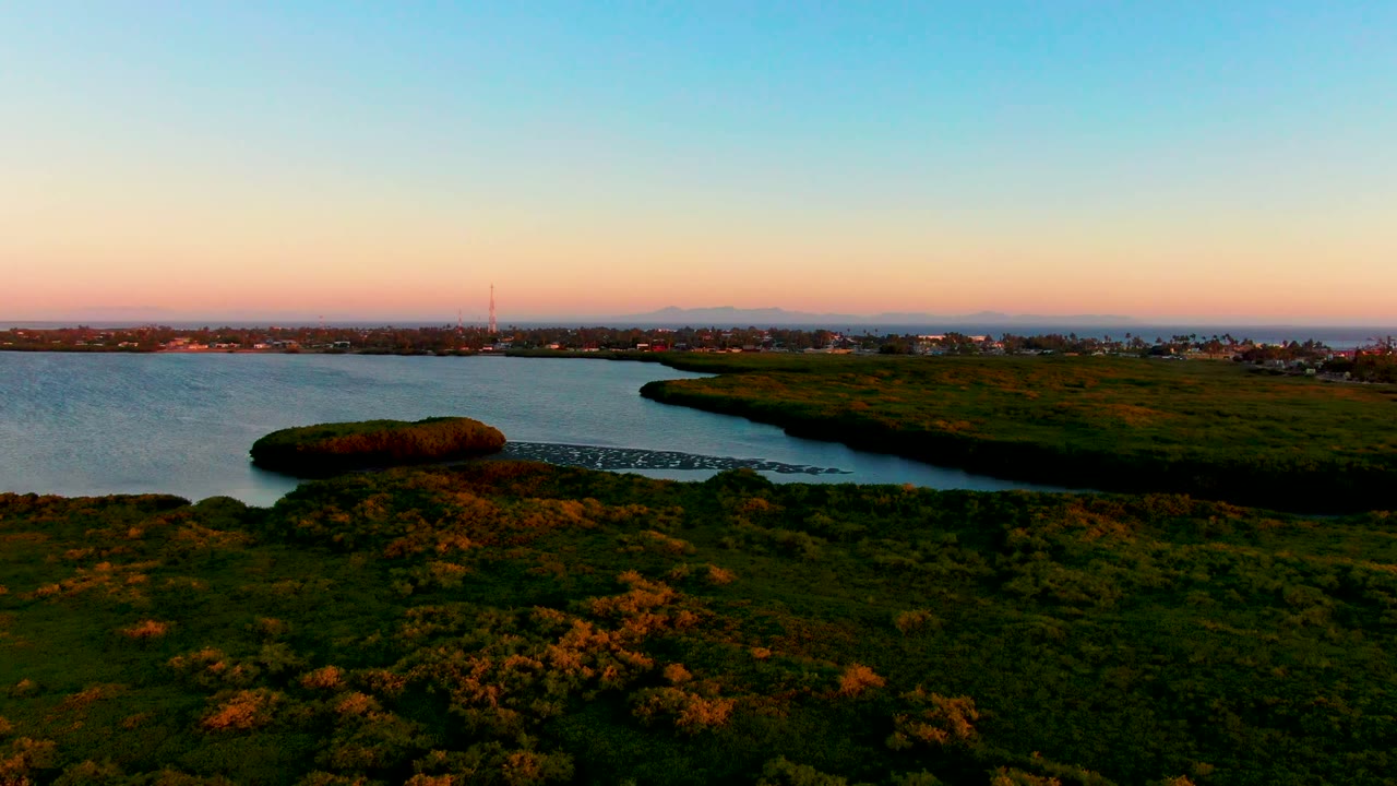 A wide aerial frame shows the San Carlos coastal inlet, grassy foreground, exposed shoreline, and a distant low skyline with no visible people.