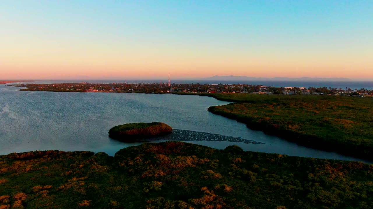 Opening aerial view in San Carlos, Baja California Sur, showing calm water, dark foreground brush, a grassy shoreline, low town buildings, a tower on the horizon, warm sunset color, and no visible people.