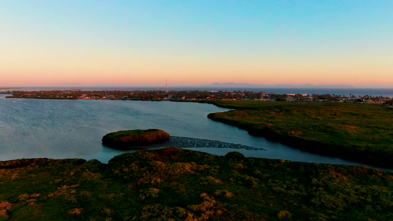 Wide coastal sunset view with water, marshy land, town buildings, and a tower near the horizon.