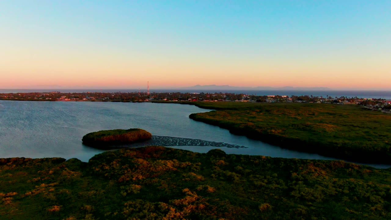 A later aerial frame in San Carlos, Baja California Sur, showing the same calm water and green shoreline with the coastal town stretched along the horizon, orange light near the skyline, and no visible people.