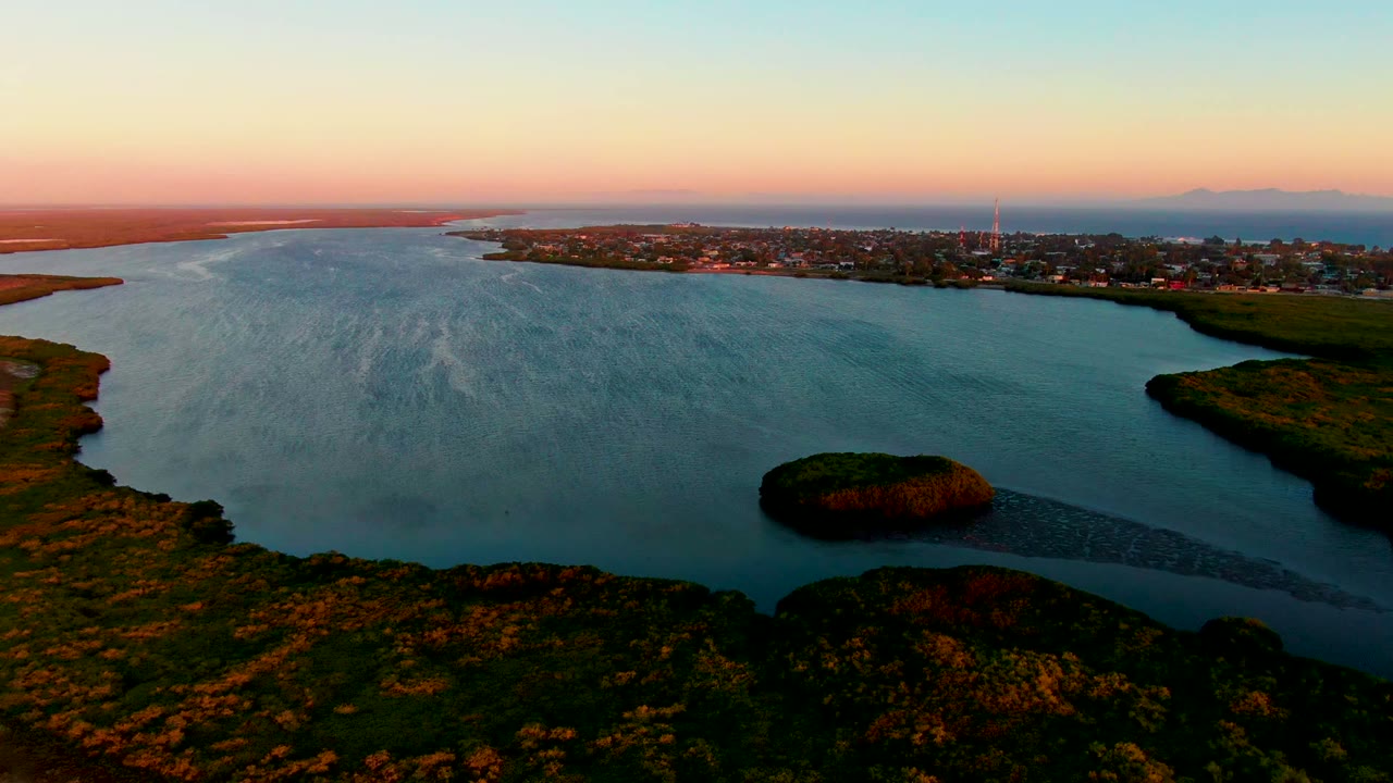 Aerial view over San Carlos, Baja California Sur, showing blue bay water, a small island near the lower center, green coastal vegetation, a distant town, pink sky, and no visible people.