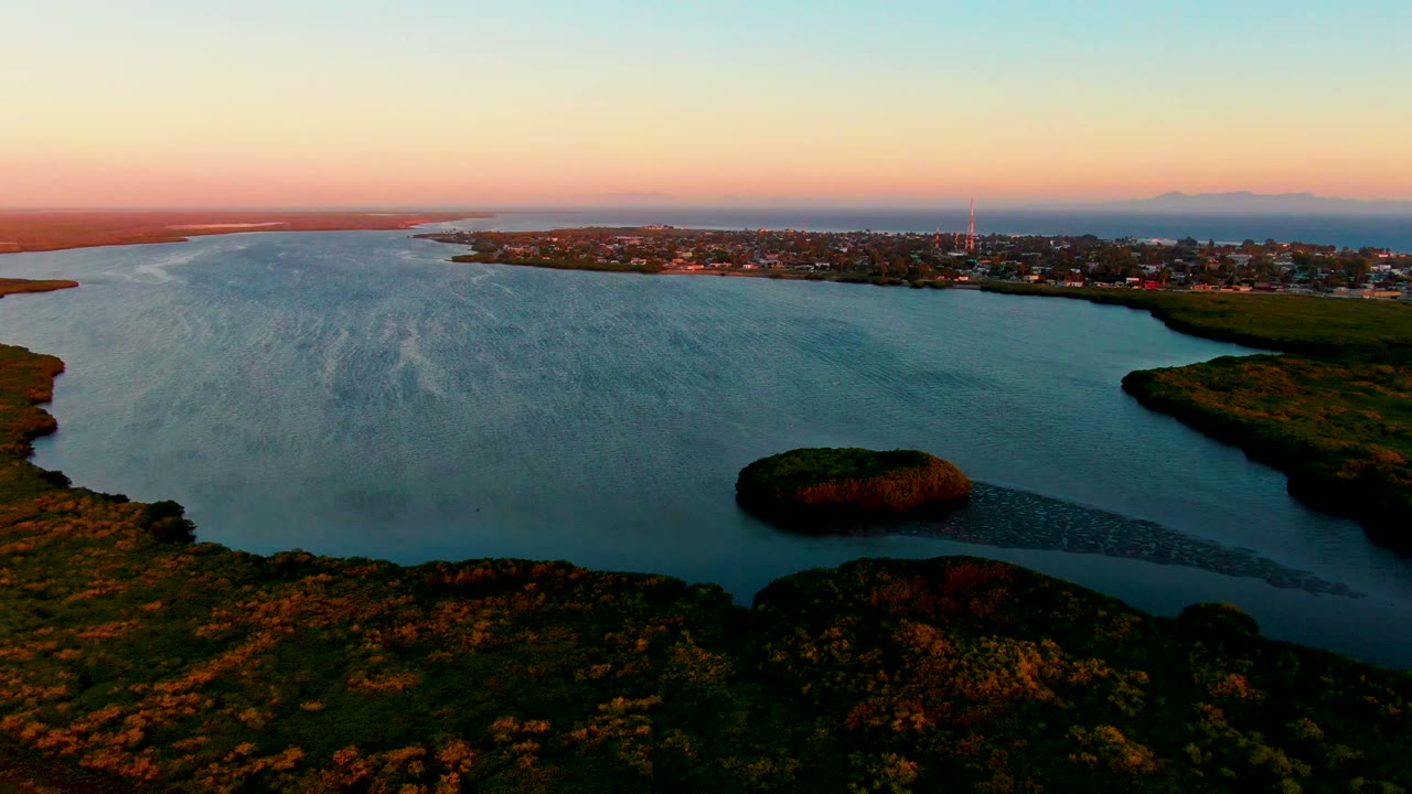 Wide aerial bay view with island, shoreline town, green land, and pastel sky.
