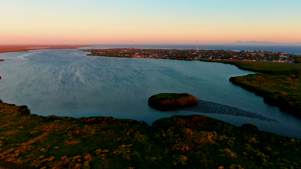 Drone-style view over San Carlos, Baja California Sur, with blue water filling the left side, the small island near the lower middle, marshy green shoreline at right, a far town, and no visible people.
