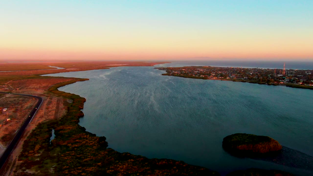 Wide aerial view in San Carlos, Baja California Sur showing a blue-green river, a small island near the lower right, dry shoreline with a road at left, distant town, sunset gradient sky, and no visible people.