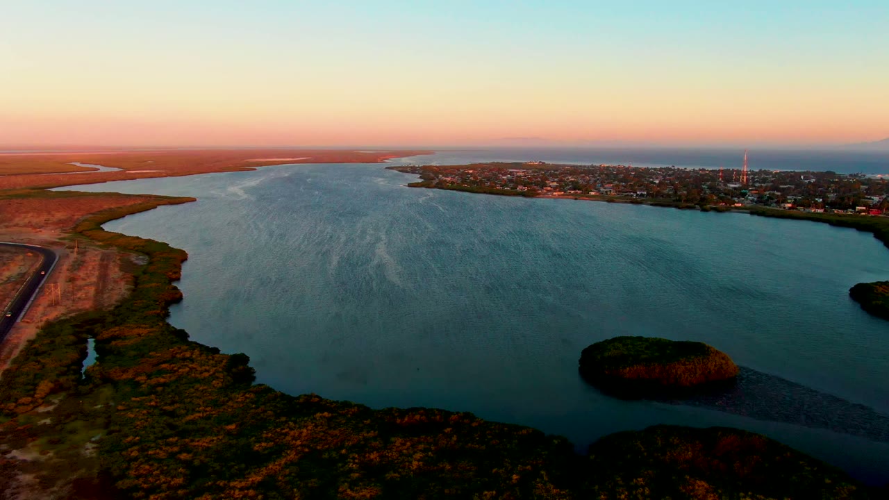 San Carlos aerial view showing the river channel stretching toward the horizon, a road along the dry left shoreline, a reed-covered island near the lower right, town buildings in the distance, and no visible people.
