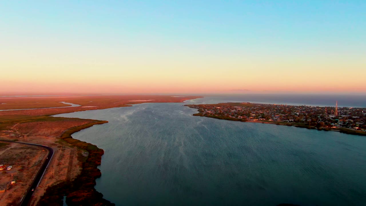 Opening aerial frame over San Carlos, Baja California Sur, with broad estuary water between dry left shoreline and a coastal town on the right, no visible people.