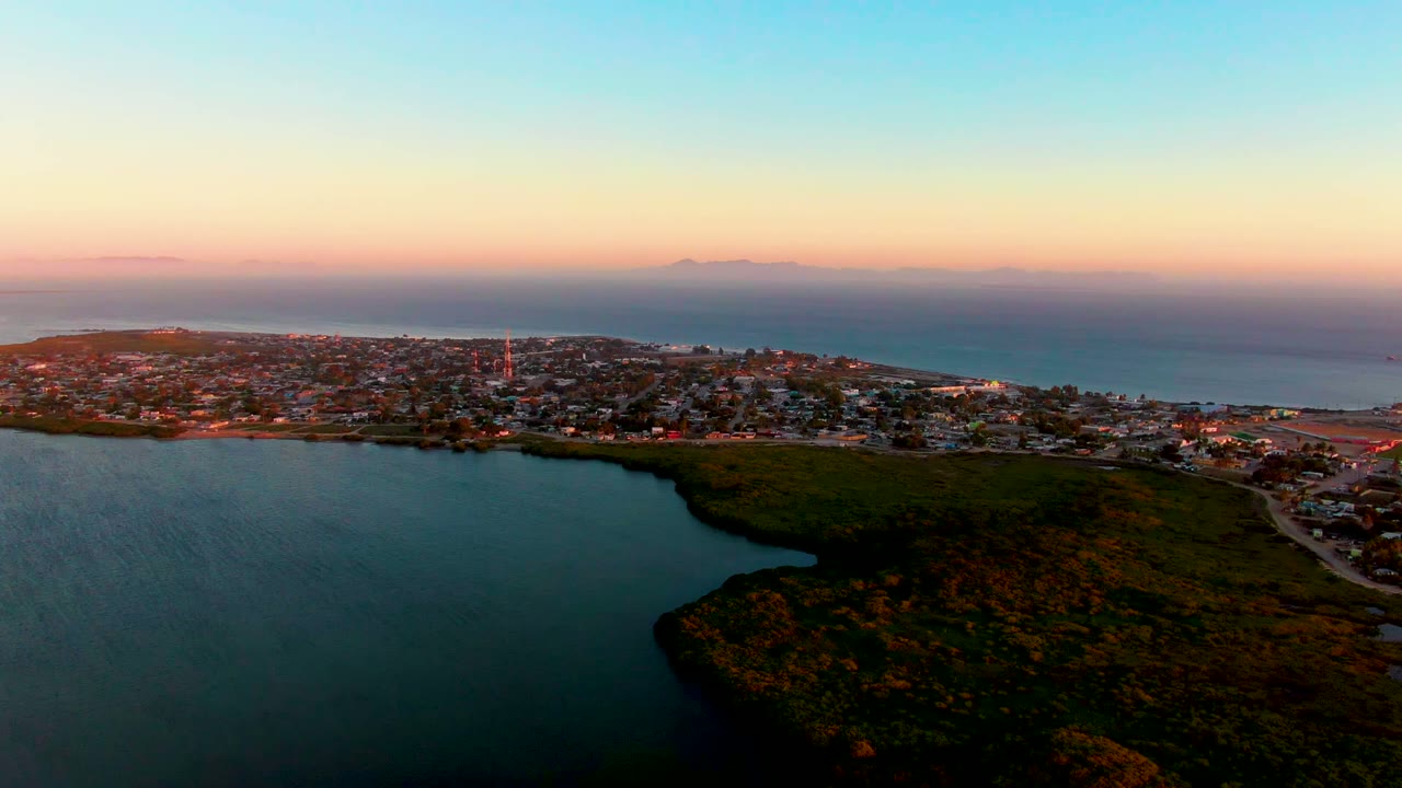 Aerial view of San Carlos, Baja California Sur at dusk, with a dark lagoon in the foreground, a green hill, the coastal town, the ocean horizon, and no visible people.