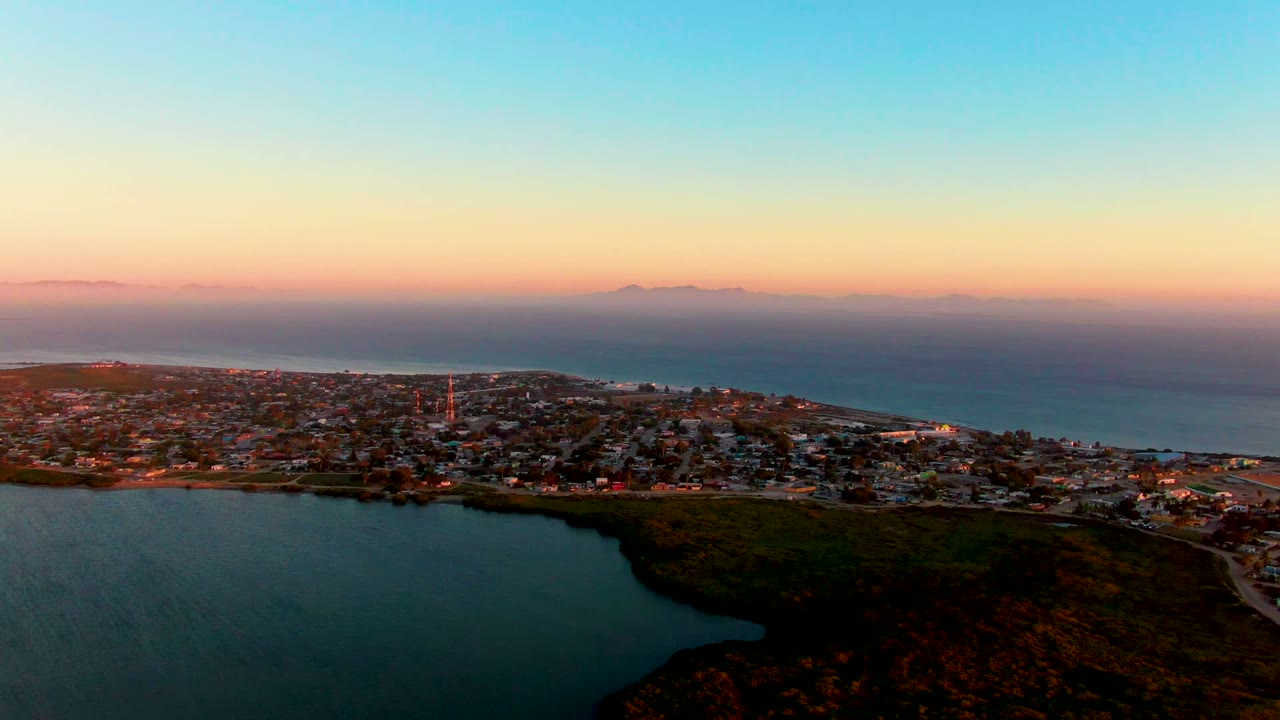 Wide drone view of lagoon water beside a coastal town under a pastel dusk sky.