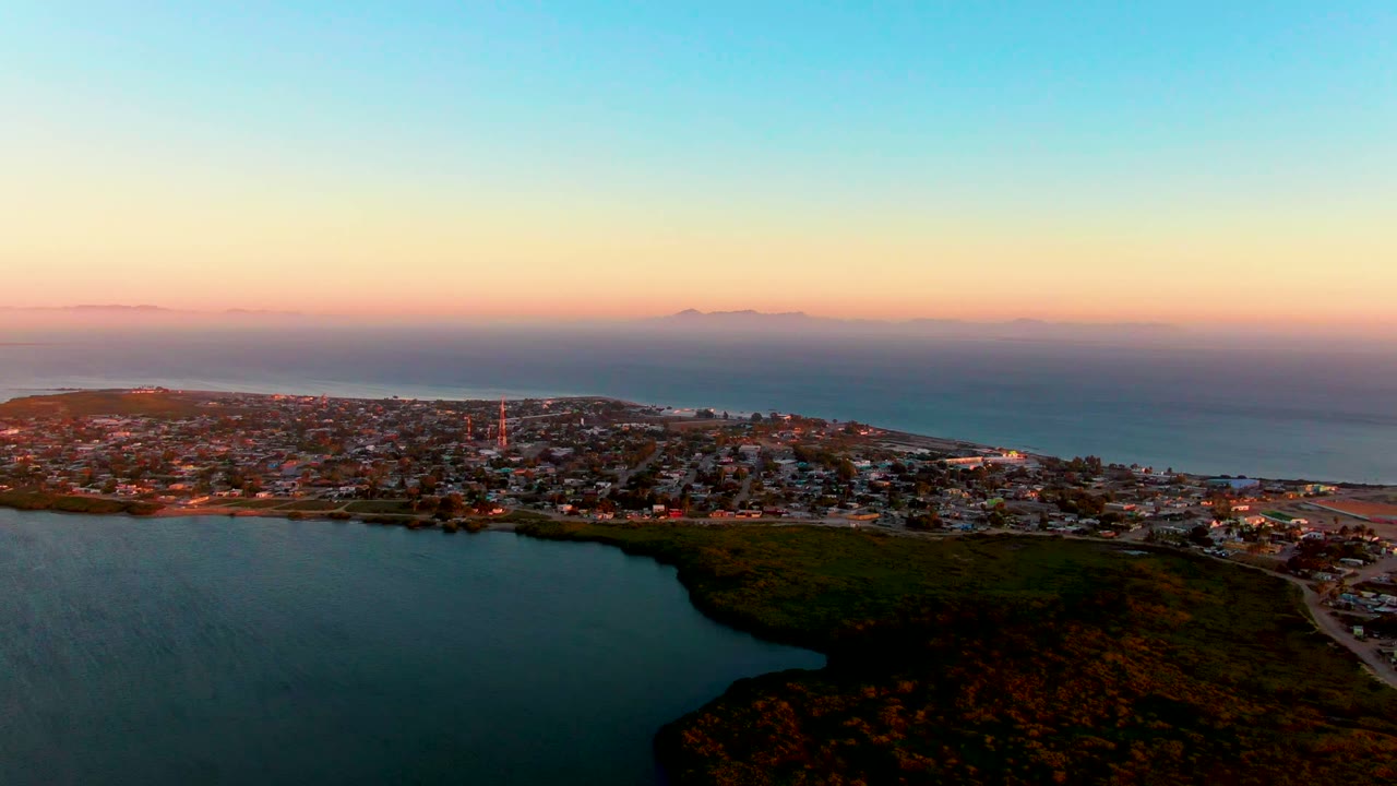 Drone-height view of San Carlos at dusk with the lagoon curving through the foreground, town structures along the shore, ocean beyond, and no visible people.