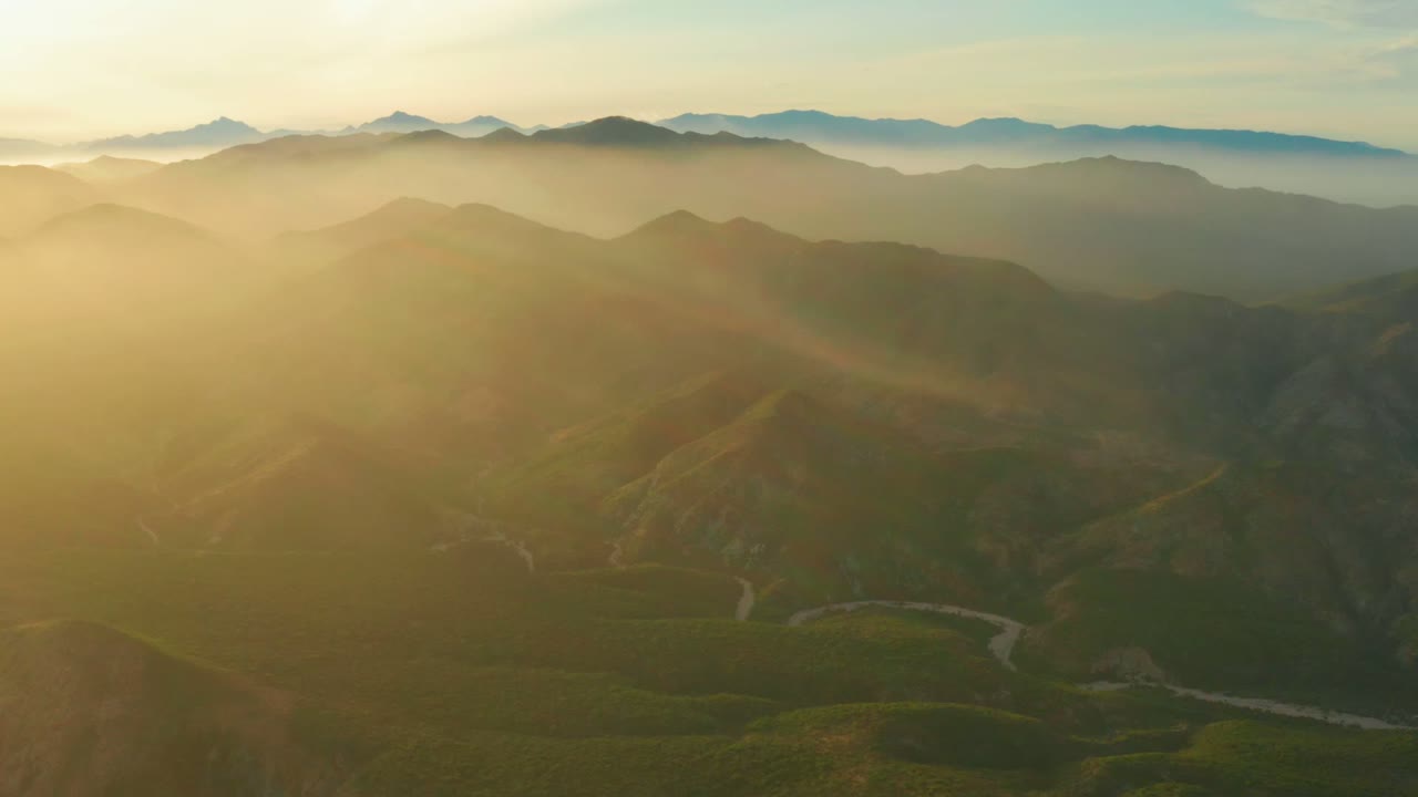 Wide aerial view of layered mountain valleys near Cabo Pulmo, Baja California Sur, with golden haze entering from the left, a winding pale road or wash in the lower valley, and no visible people.