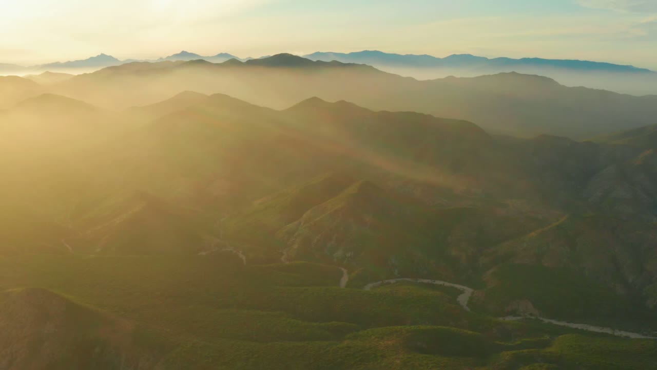 Wide drone view near Cabo Pulmo showing darker foreground hills, misty layered mountains in the distance, golden light washing in from the left, and no visible people.