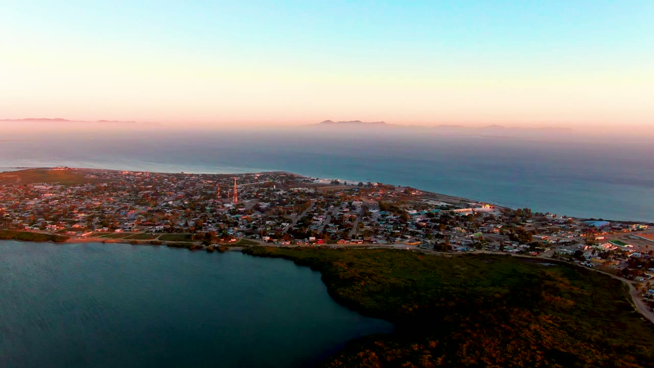 Aerial view over San Carlos, Baja California Sur at dusk, with a dark lagoon in the foreground, the town across the middle of the frame, ocean on the horizon, and no visible people.