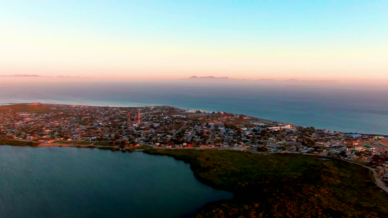 Aerial dusk view of San Carlos, Baja California Sur, showing clustered buildings, a vertical tower-like structure, lagoon water below, ocean beyond, and no visible people.