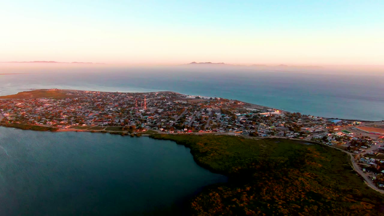 Wide aerial frame over San Carlos, Baja California Sur, with dark water and green shoreline vegetation in the foreground, town on the peninsula, distant islands on the horizon, and no visible people.