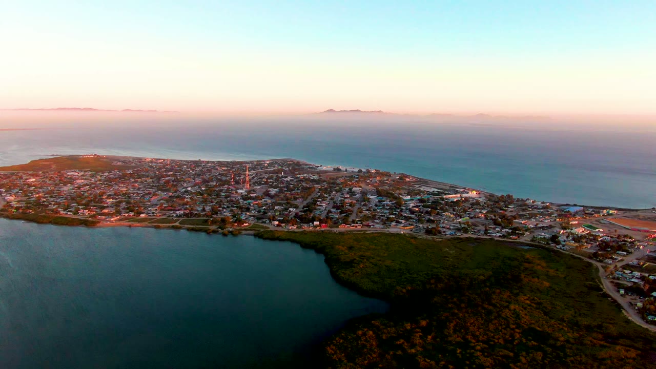 Aerial coastal town view with water in the foreground and hazy islands across the ocean horizon.