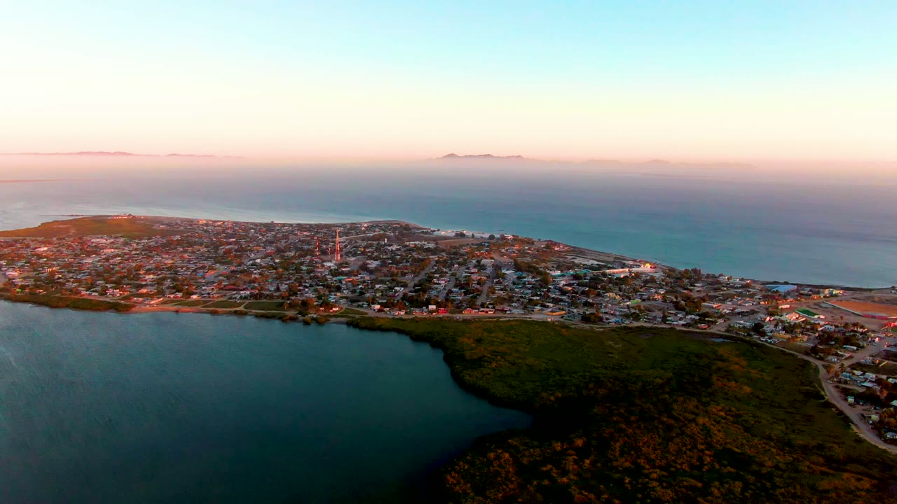 Mid-clip aerial frame over San Carlos with broad dark water below, a dense town grid across the peninsula, faint islands beyond the ocean, and no visible people.