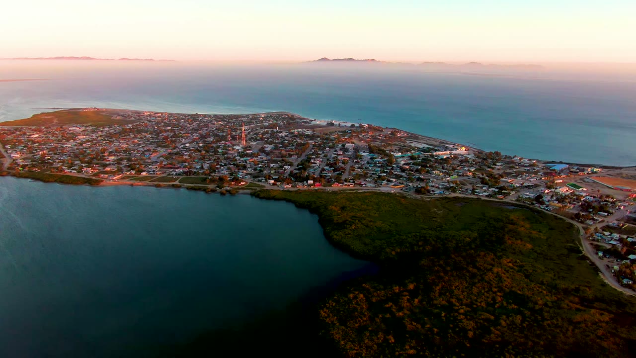 Wide aerial frame over San Carlos, Baja California Sur, showing a coastal town between a dark inlet and open ocean under a pink-blue dawn sky, with no visible people.