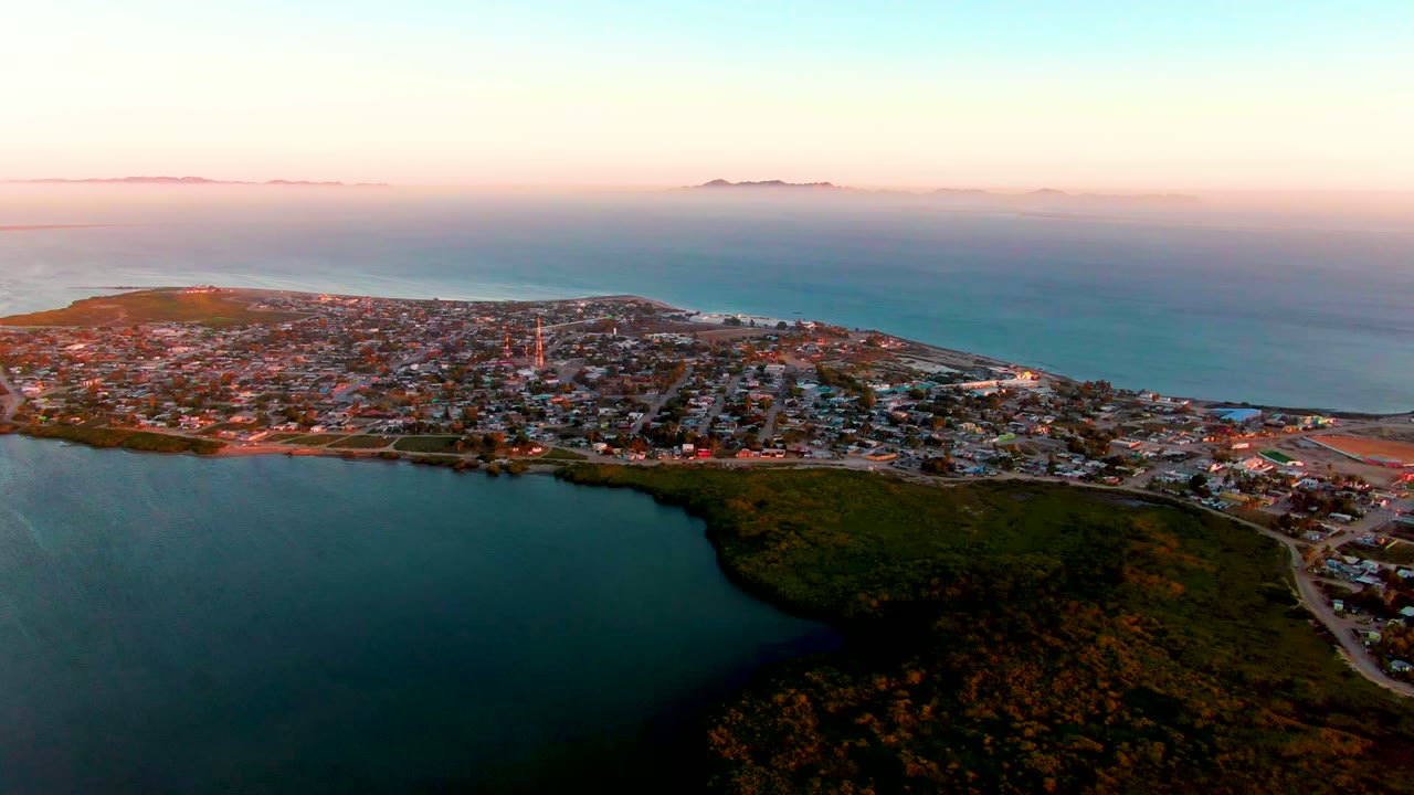 San Carlos appears in a broad aerial composition with dark inlet water in front, green hilly vegetation to the lower right, dense low buildings in the middle, and the ocean horizon behind, with no visible people.