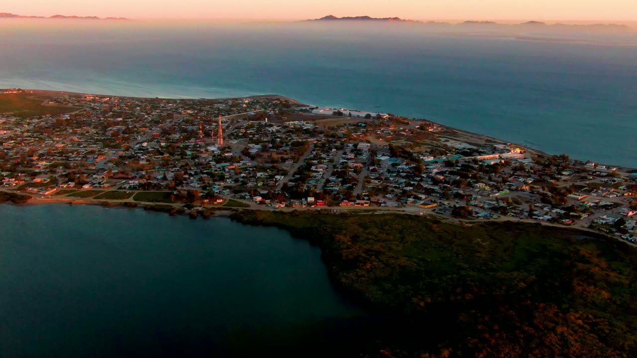 High aerial view over San Carlos, Baja California Sur, with dark bay water in the foreground, a coastal town across the middle, ocean and low mountains on the horizon, and no visible people.