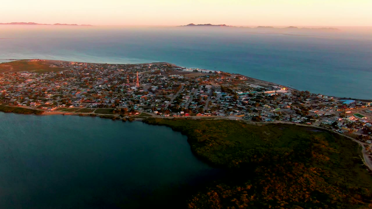 San Carlos aerial view with green shoreline vegetation bordering the bay, the town spread across the peninsula, ocean behind it, soft orange-pink light near the horizon, and no visible people.