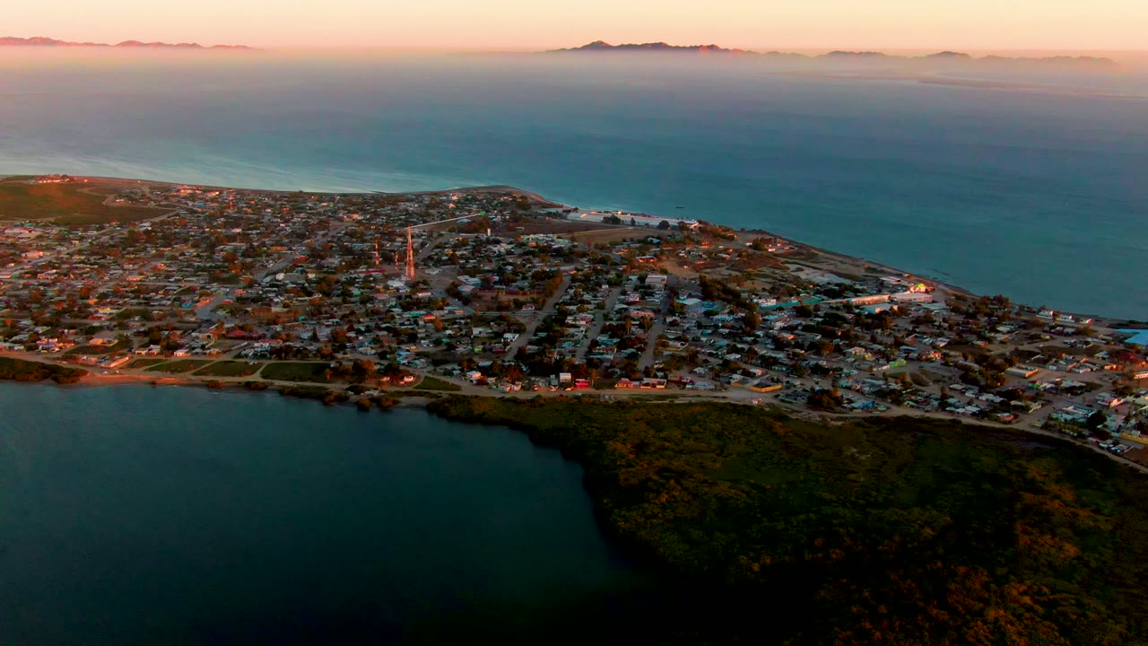 Opening frame of San Carlos, Baja California Sur from above, with dark foreground water, dense town blocks, a steeple near the center-left, pink dusk sky, distant hills, and no visible people.
