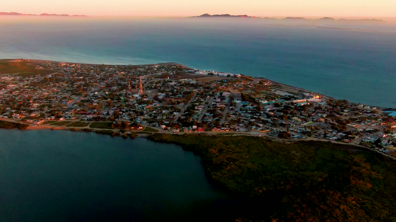 Third frame from the aerial pass over San Carlos, Baja California Sur, with the town stretching along the shore, a tall steeple visible among the buildings, dark green land at the lower right, and no visible people.
